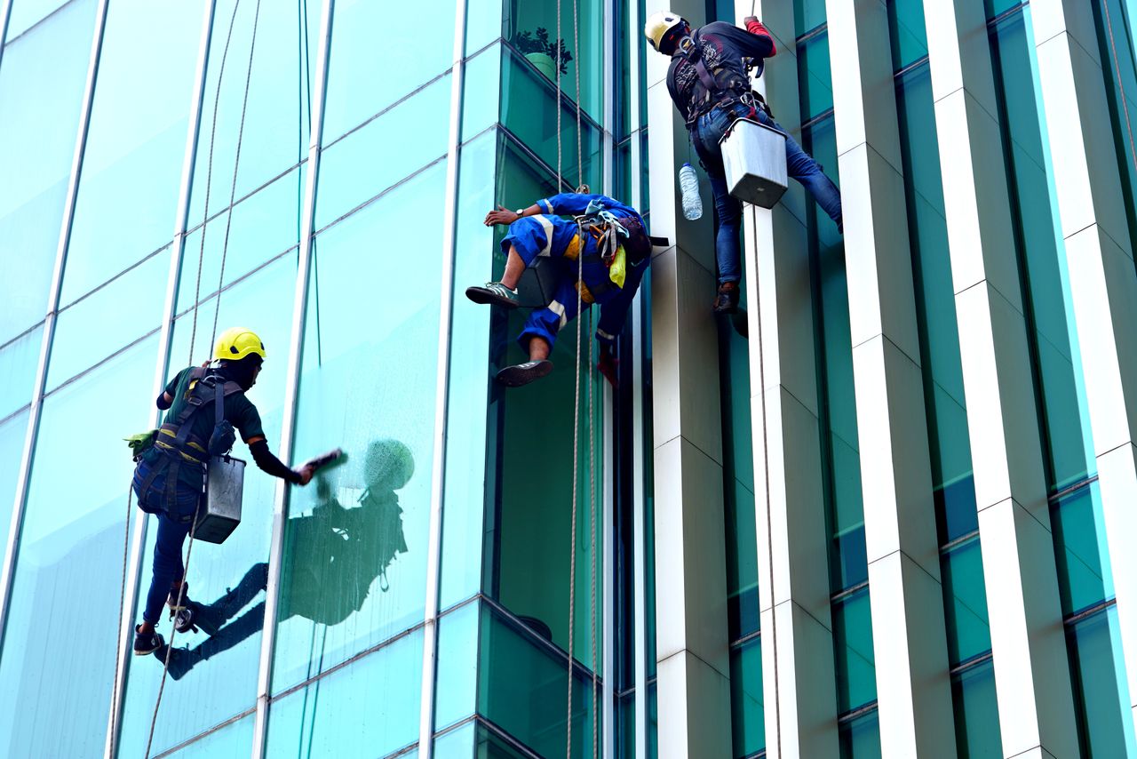 services-04 During the day, three window cleaners suspended from ropes are cleaning the facade of a modern glass building.